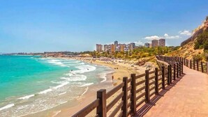 Un paseo marítimo con baranda de madera que conduce a una playa de aguas turquesas cristalinas y un paisaje urbano al fondo.