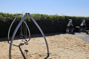 Een schommel in een zandbak, met een haag op de achtergrond.