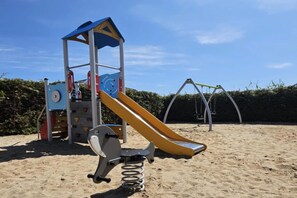 Une aire de jeux avec un toboggan, des balançoires et des appareils de musculation, le tout installé sur un sol sablonneux.