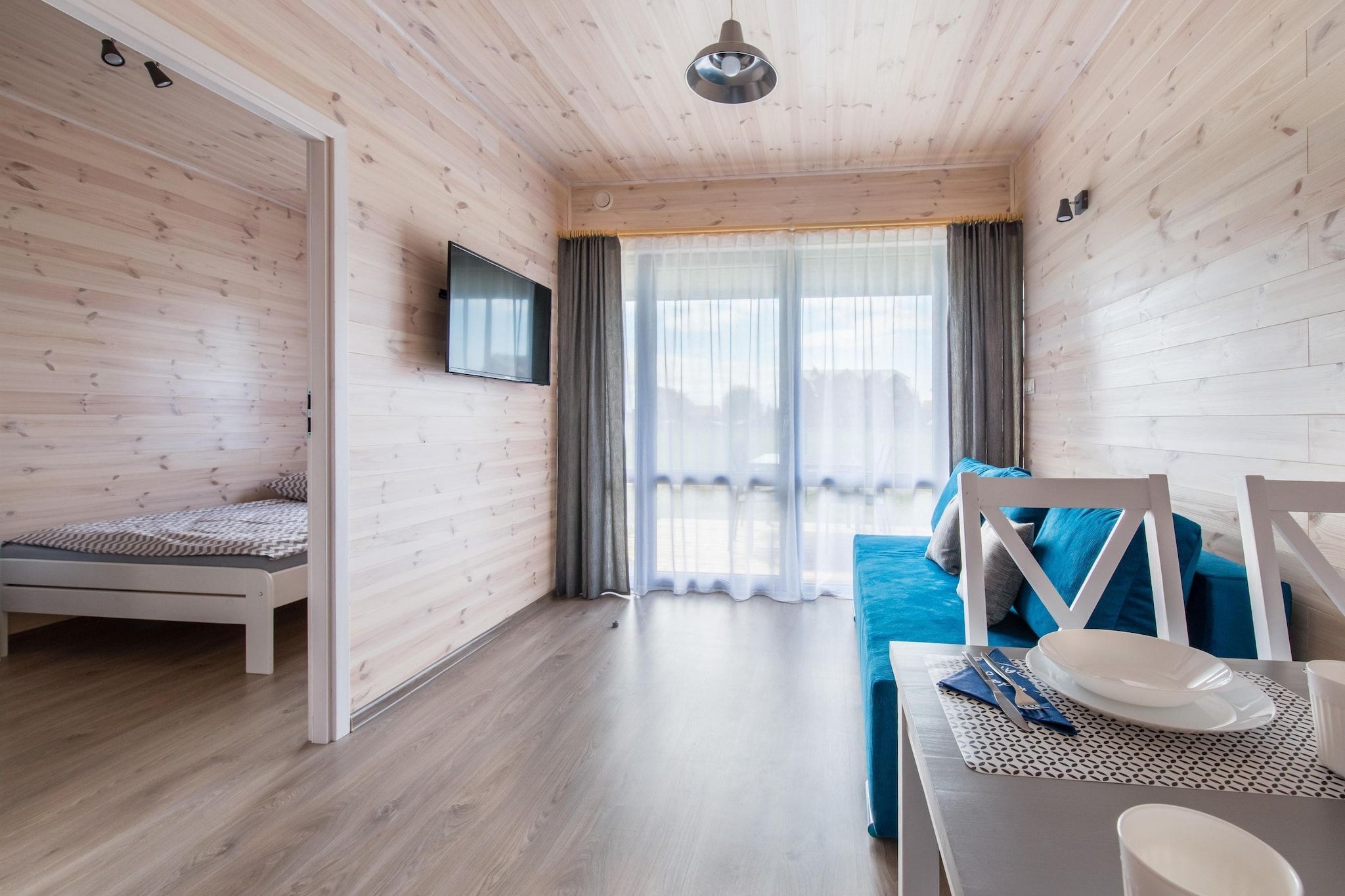 A room with a bed, a sofa, a dining table with a plate and cutlery, a TV on the wall, and a window with curtains.