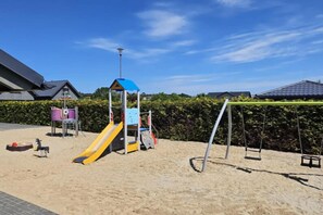 Un parque infantil con tobogán y columpios, rodeado de arena y follaje.