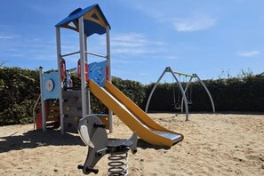 Ein Spielplatz mit Rutsche und Schaukeln, umgeben von Sand und einem Hecken