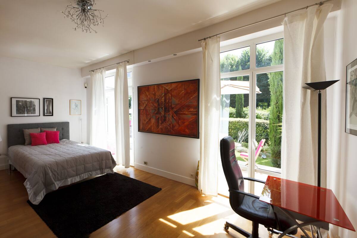 A bedroom with a bed, a desk, and a chair. There is a window with curtains and a view of greenery outside.