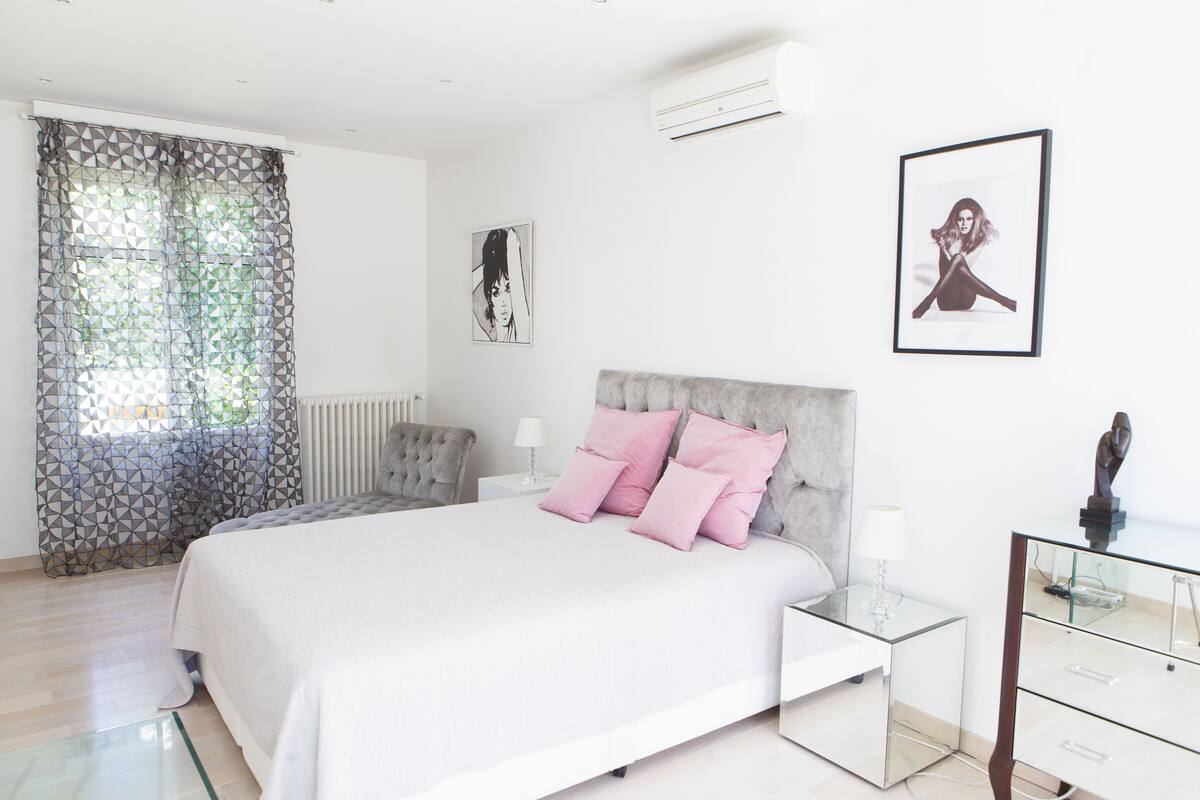 A modern bedroom with a large bed, a glass nightstand, a grey upholstered headboard, and a white radiator.