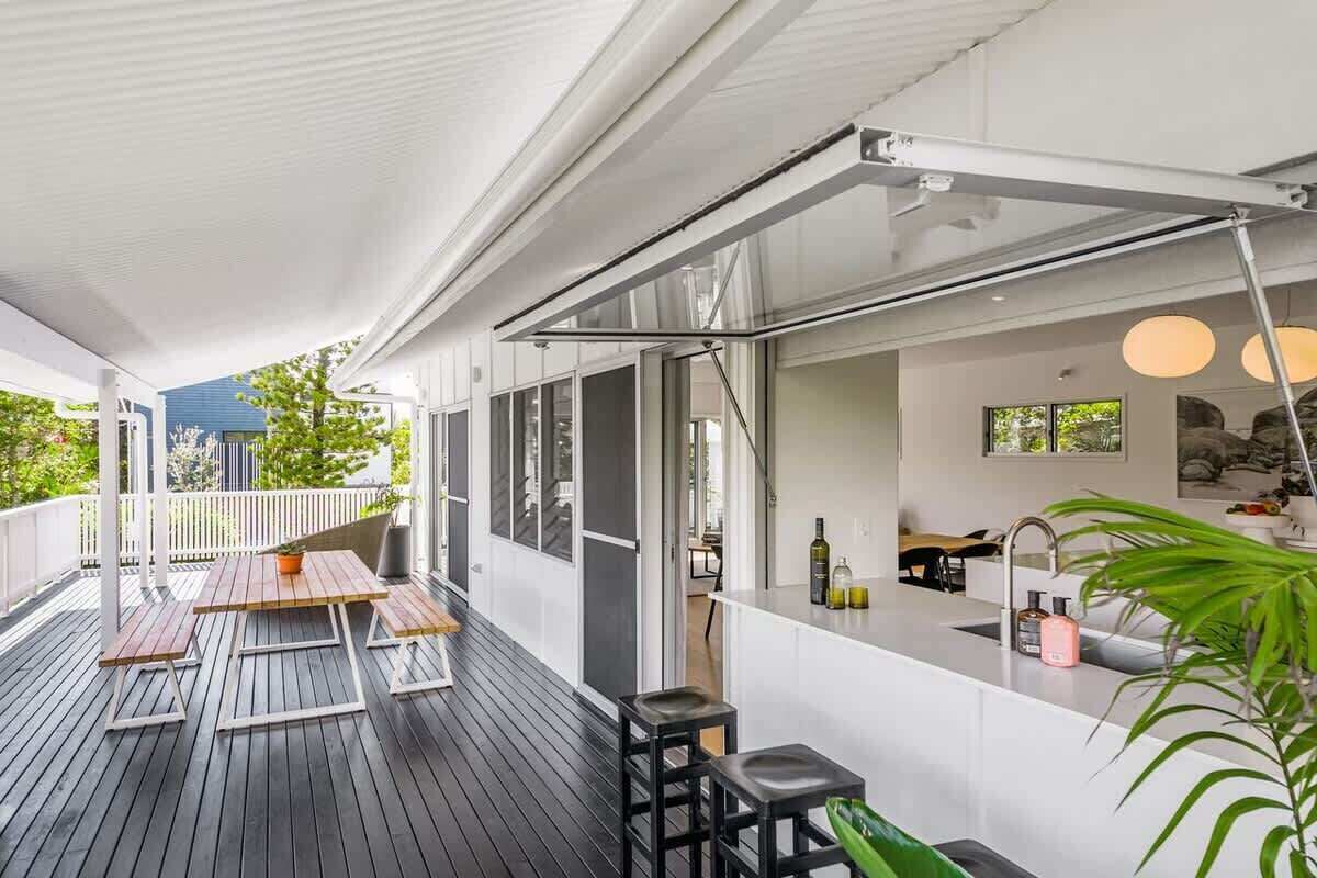 Een overdekt buitengebied met een houten terras, een bar inclusief krukken, en uitzicht op de groene omgeving.