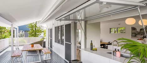 Een overdekt buitengebied met een houten terras, een bar inclusief krukken, en uitzicht op de groene omgeving.