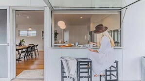 A modern kitchen with a dining area visible through an open door, a person reading a magazine, and a bar stool. - Ukiyo (Cabarita Beach)