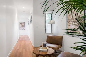 A hallway with a wooden floor, a brown leather sofa, a round table with books and a glass object, and a large plant. - Ukiyo (Cabarita Beach)