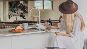A person reading a magazine at a kitchen counter with a bread basket and a bottle of wine.
