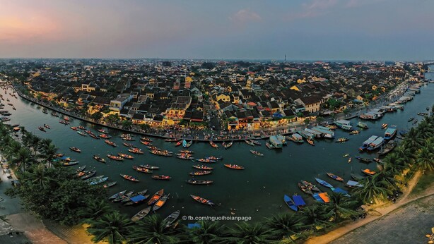 An aerial view of a riverside town with numerous boats docked along the waterway, surrounded by traditional buildings and palm trees. - LA GAI HOMESTAY HOI AN (Da Nang)