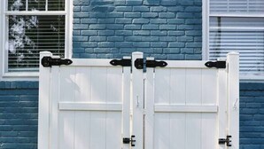 A white gate with black hinges in front of a blue brick house.