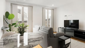 A modern living room with a grey sofa, a marble coffee table, a TV on a black stand, and large windows offering a view of the city. - GuestReady - Trendy getaway in the heart of Porto (Porto)