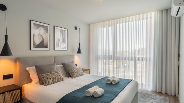 Habitación de hotel moderna con una cama grande, dos mesitas de noche, aire acondicionado de pared y un amplio ventanal con cortinas traslúcidas.