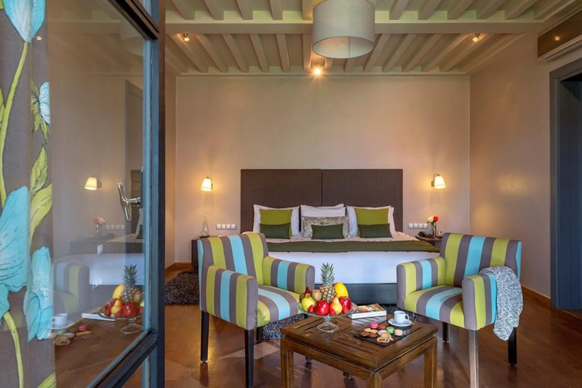A hotel room with a bed, two striped armchairs, a coffee table with fruit, and a window with a view.