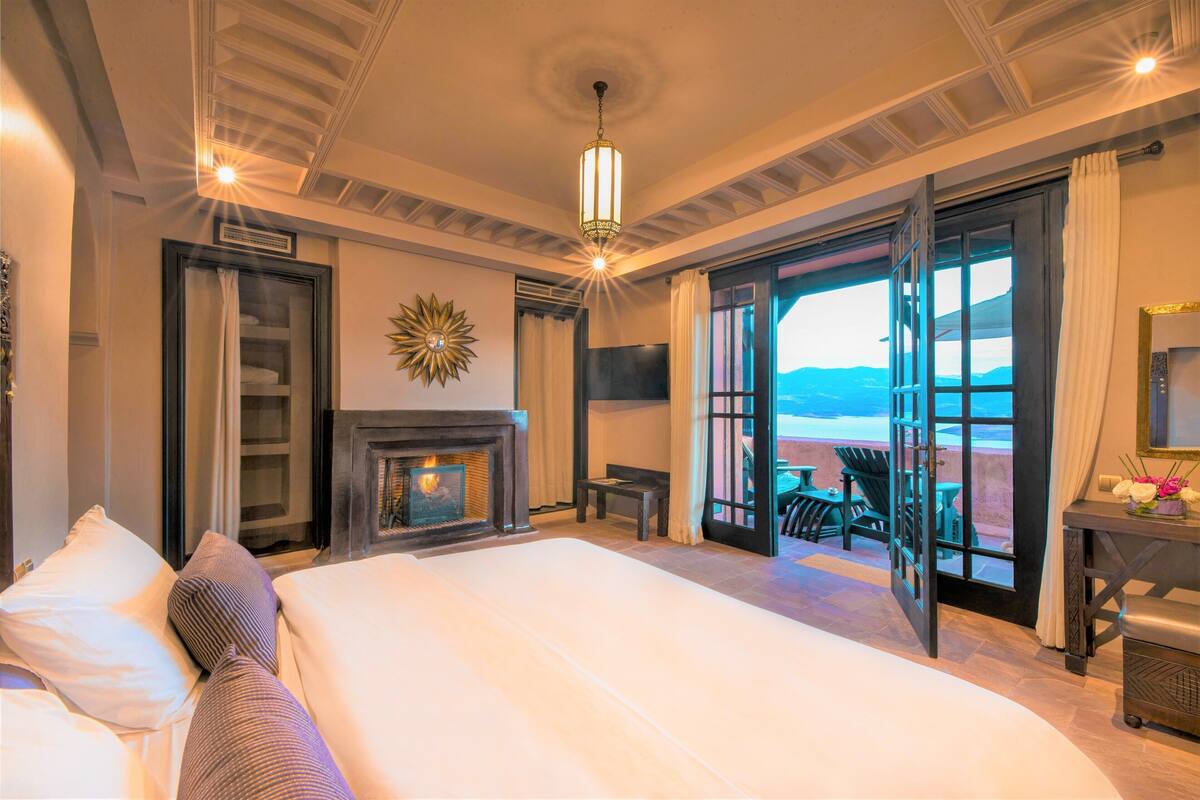 Un dormitorio amplio con una cama grande, una chimenea, un balcón con mobiliario exterior y vistas a las montañas.