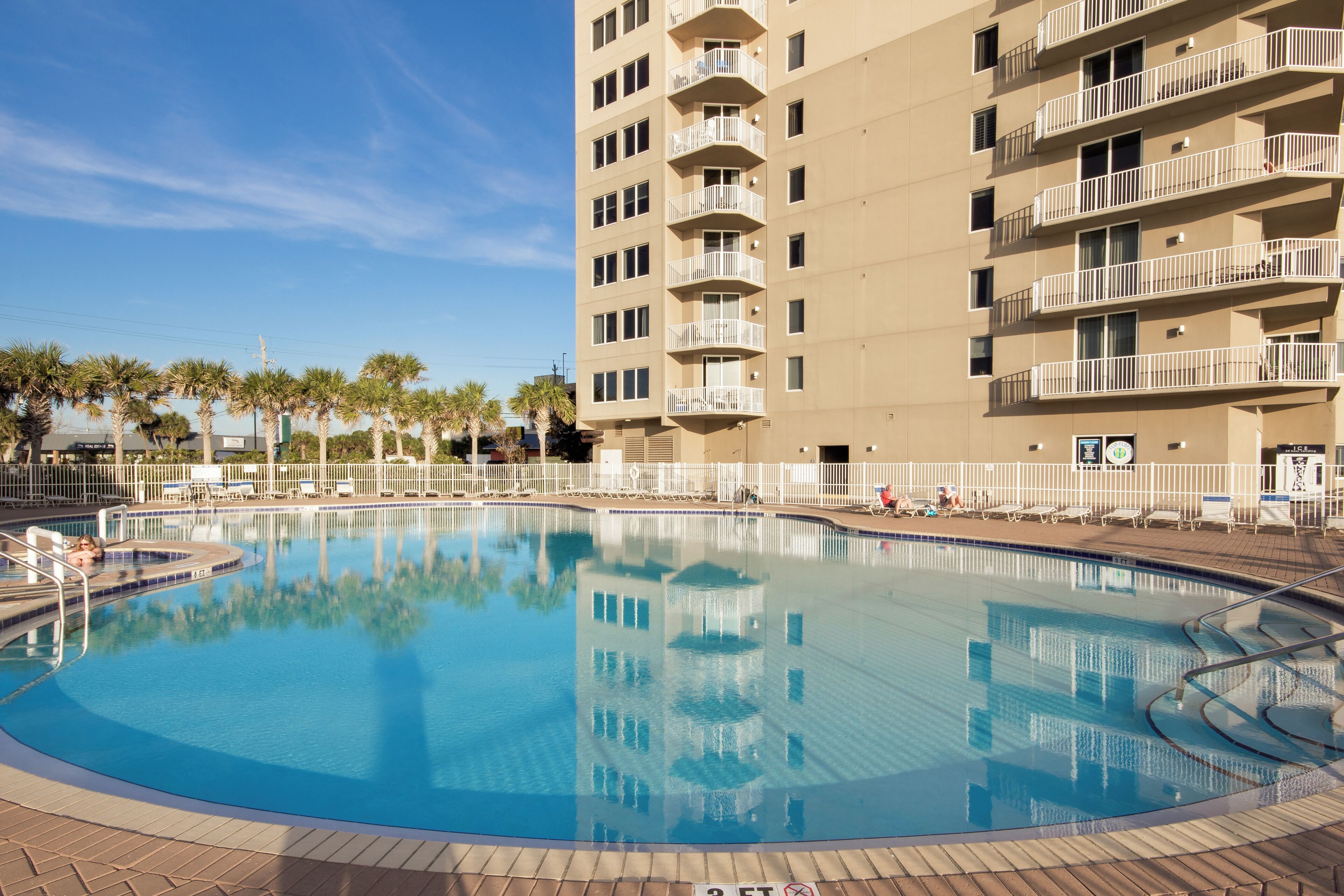 A swimming pool with a clear blue water surface, surrounded by a white fence and palm trees.