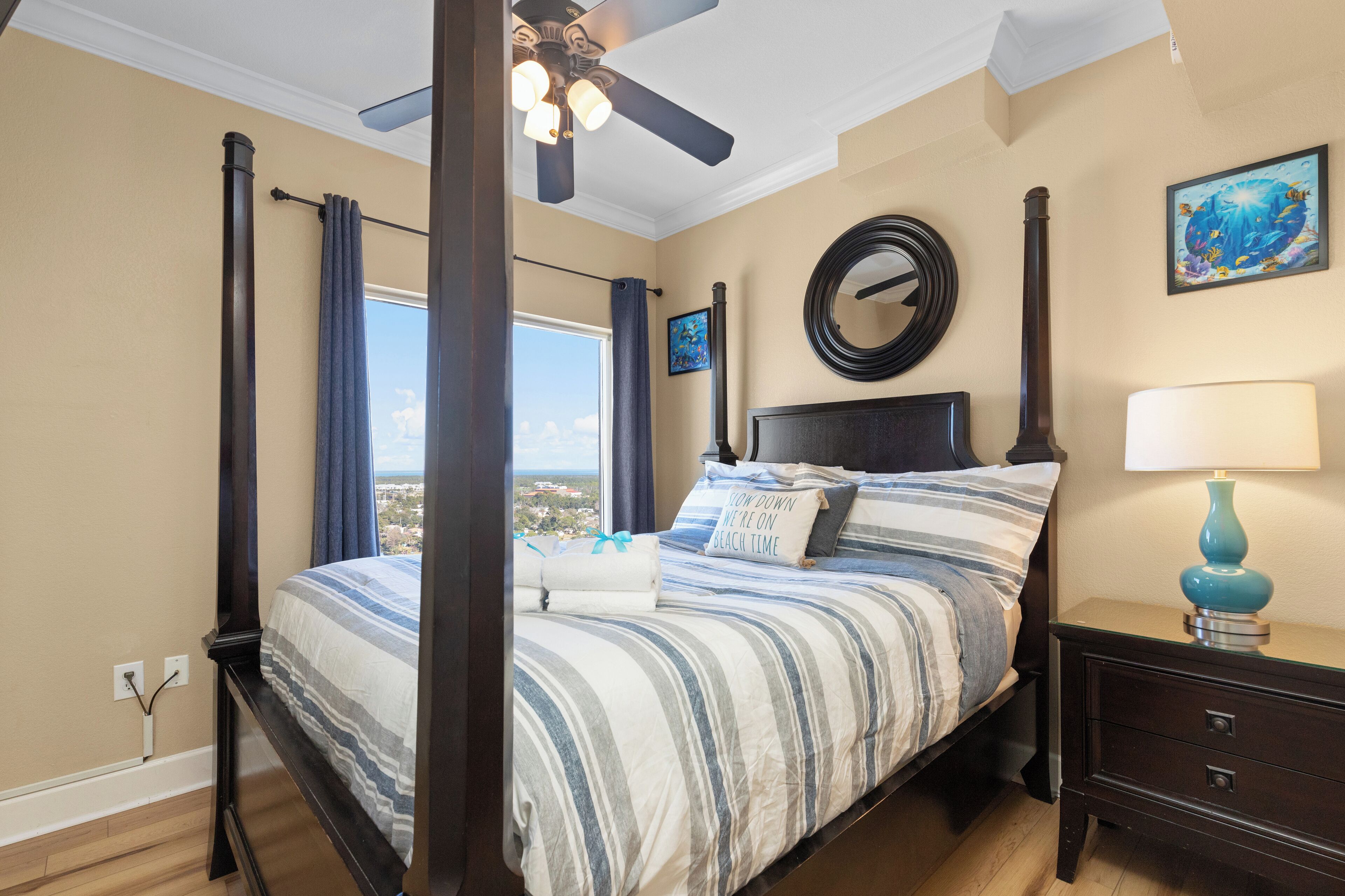 Un dormitorio con cama con dosel, ventilador de techo, vista de la ciudad y mesita de noche con lámpara.