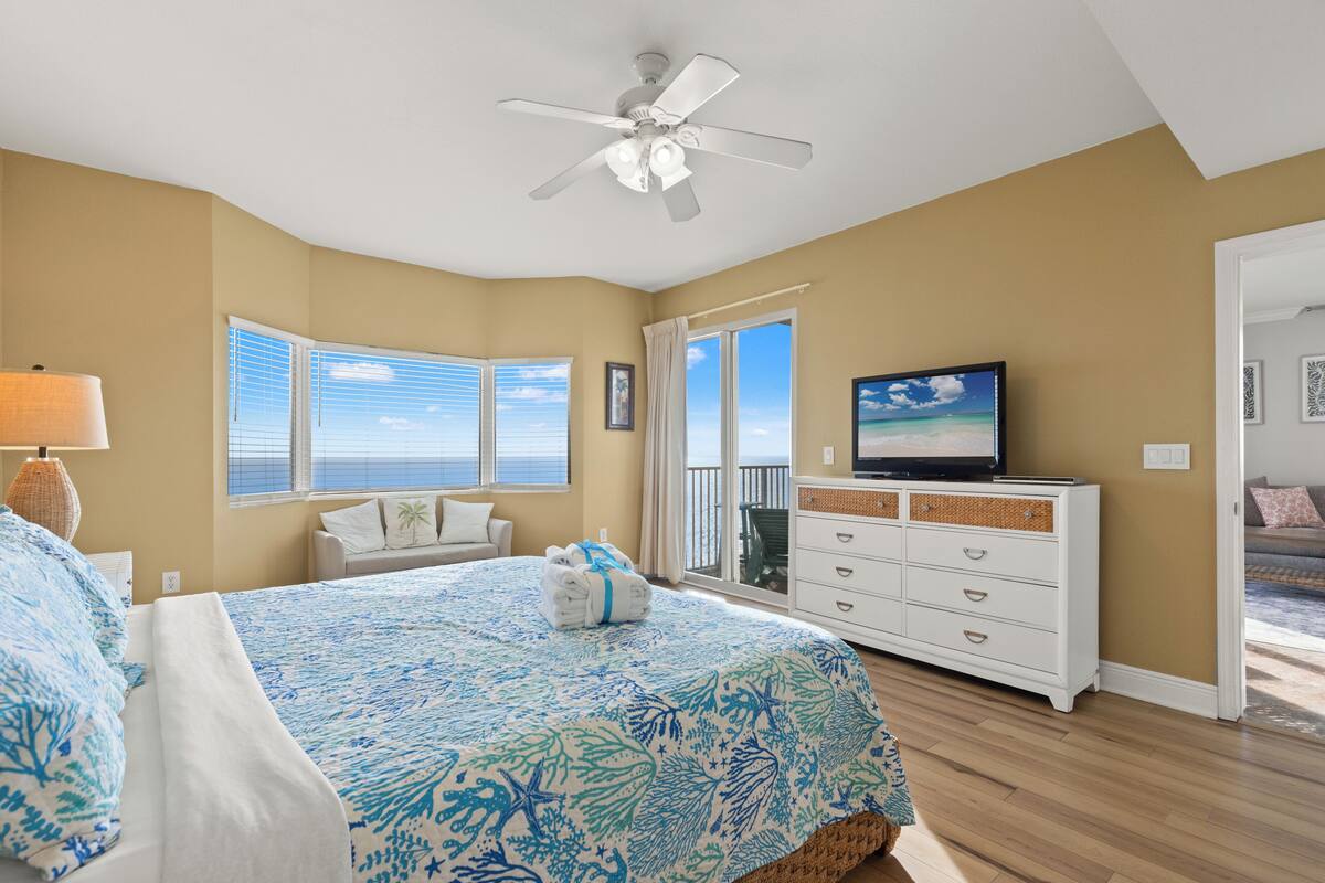 Een slaapkamer met een bed, een plafondventilator, een televisie, een dressoir en uitzicht op zee.