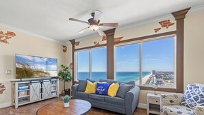 Ein Wohnzimmer mit großem Fenster mit Blick auf den Strand, einem Deckenventilator, einem grauen Sofa mit gelben Kissen, einem Holztisch und einem Beistelltisch mit einem dekorativen Gegenstand.
