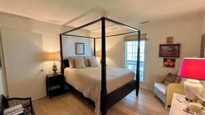 A four-poster bed with a quilt, a nightstand with a lamp, a wicker chair, and a table with a vase.