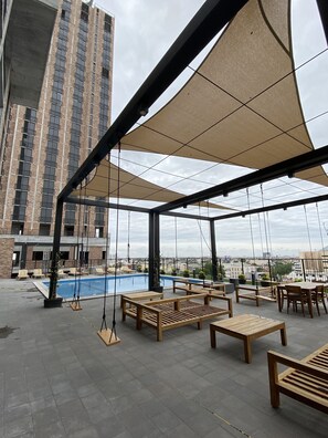 An outdoor seating area with wooden furniture and a pool, surrounded by a shaded structure and a view of a tall building.