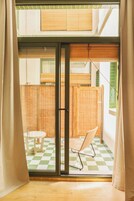 Quarto com porta de vidro deslizante para uma varanda com chão xadrez, uma cadeira de vime e uma pequena mesa.