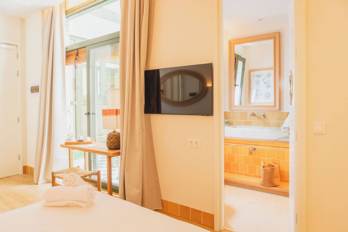 Ein Hotelzimmer mit Bett, einem an der Wand befestigten Fernseher, einem Badezimmer mit Waschbecken und Spiegel sowie einem Fenster mit Vorhängen.