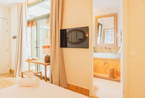 Habitación de hotel con cama, televisor montado en la pared, baño con lavamanos y espejo, y ventana con cortinas.