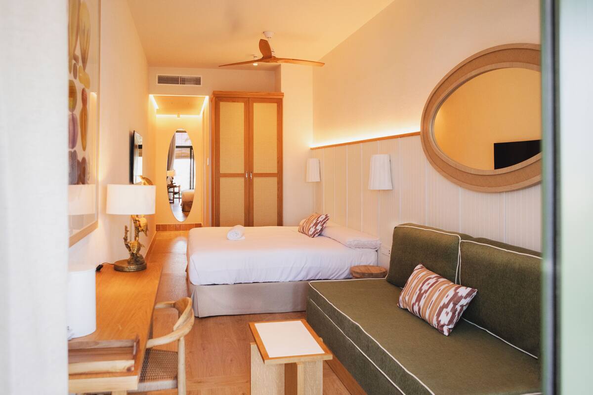 Ein Hotelzimmer mit Bett, grünem Sofa, Schreibtisch mit Lampe, Spiegel und Deckenventilator.