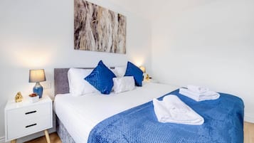 A neatly made bed with a blue blanket and white pillows, a small bedside table with a lamp, and a wall-mounted painting.