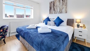 A bedroom with a large bed, a window with blinds, a radiator, a chair, and a nightstand with a lamp.