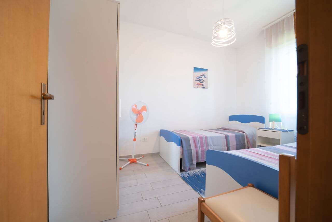 Une chambre avec deux lits, un ventilateur, une lampe et un tableau accroché au mur.