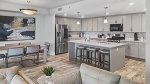 A modern kitchen with stainless steel appliances, a central island with bar stools, and a dining area with a large painting. - Blu 200 - Large 4Bd Beachfront - Great Amenities Including Beach Service! (Fort Walton Beach)