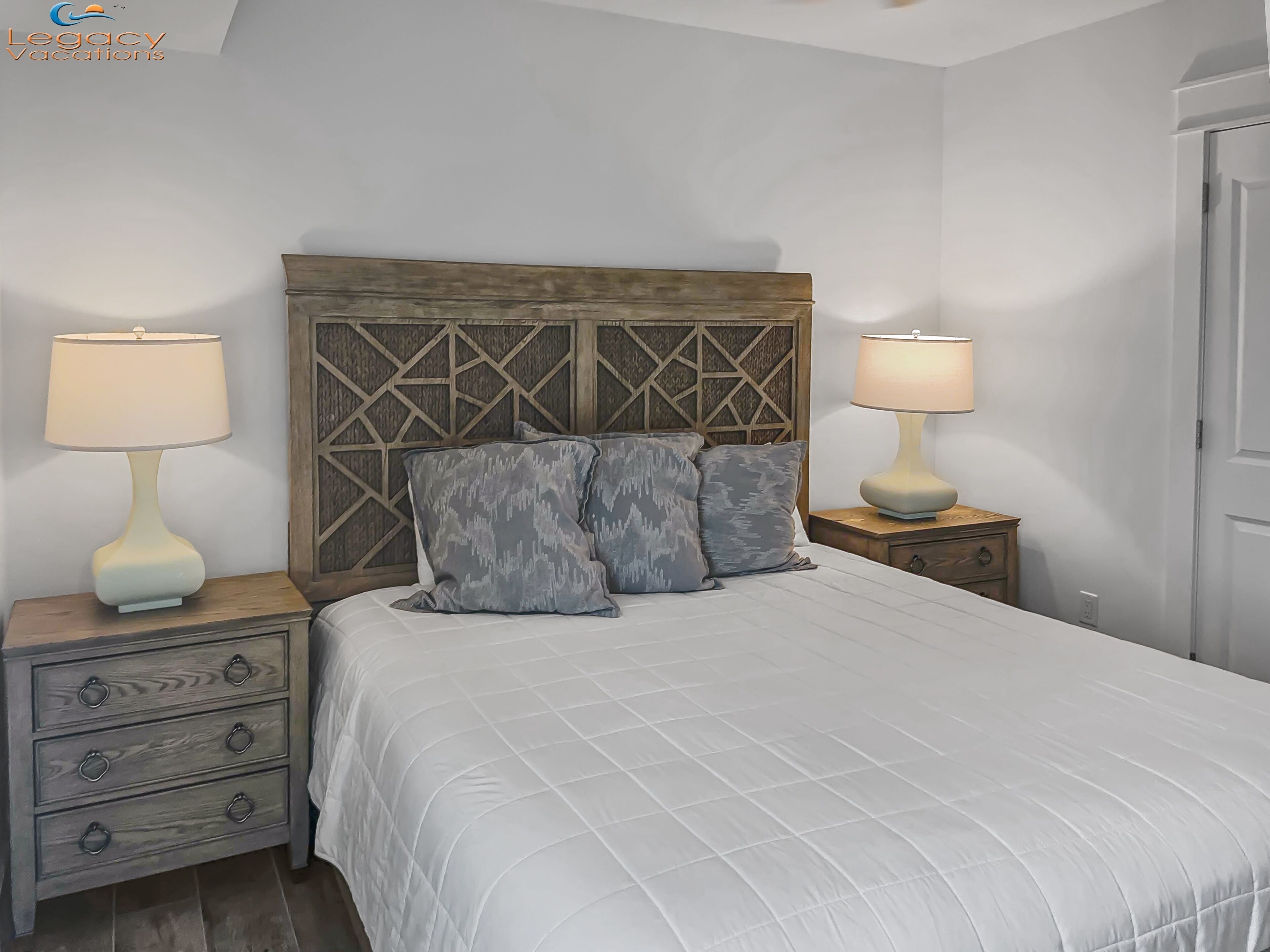 A bedroom with a wooden headboard, two bedside tables with lamps, a bed with a white quilted bedspread, and three gray patterned pillows.
