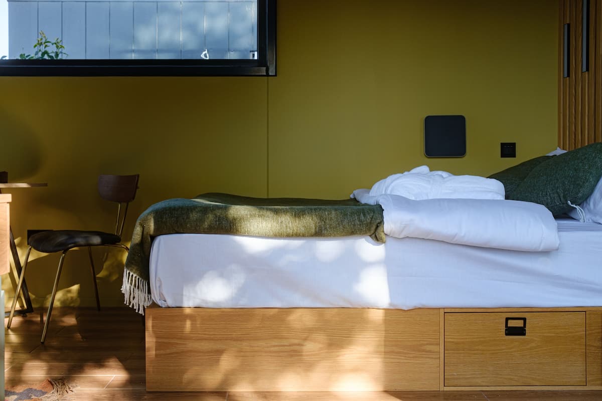 A neatly made bed with a green throw blanket and white linens, a wooden nightstand with a lamp, and a chair.