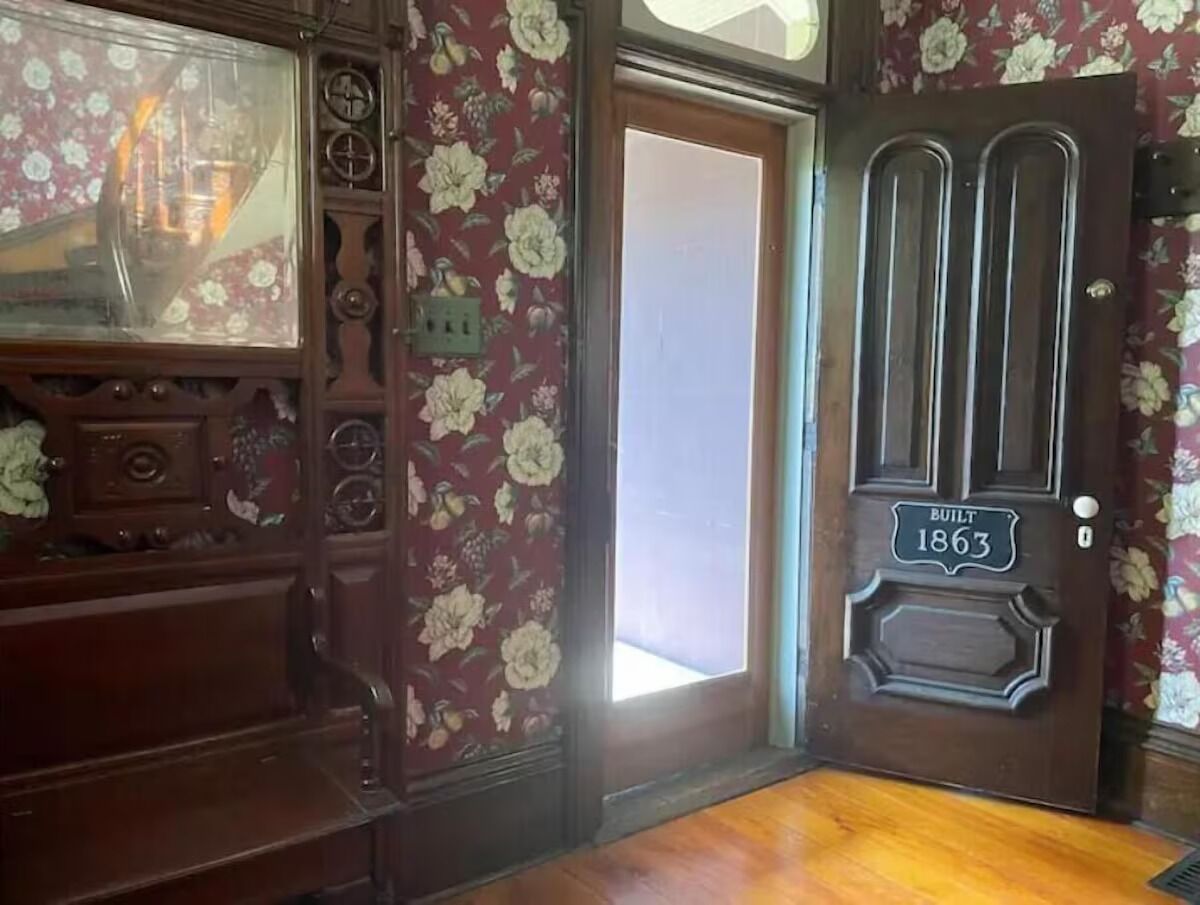 A room with floral wallpaper, a wooden door with a plaque reading "BUILT 1863", and a vintage radio.