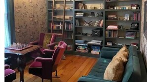 A cozy living room with a wooden floor, a green sofa, a chess table, and a bookshelf filled with books and decorative items.