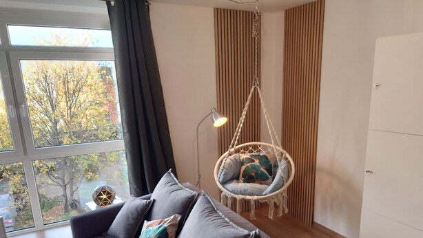 Een moderne woonkamer met een grijze bank, een hangstoel met kussens, een vloerlamp en uitzicht op bomen door het raam.