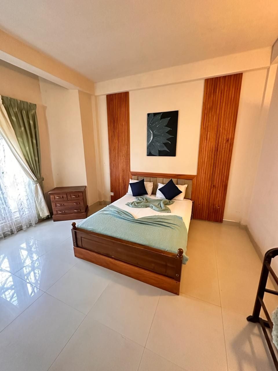 A bedroom with a wooden bed, a nightstand, a wall-mounted artwork, and a window with curtains.