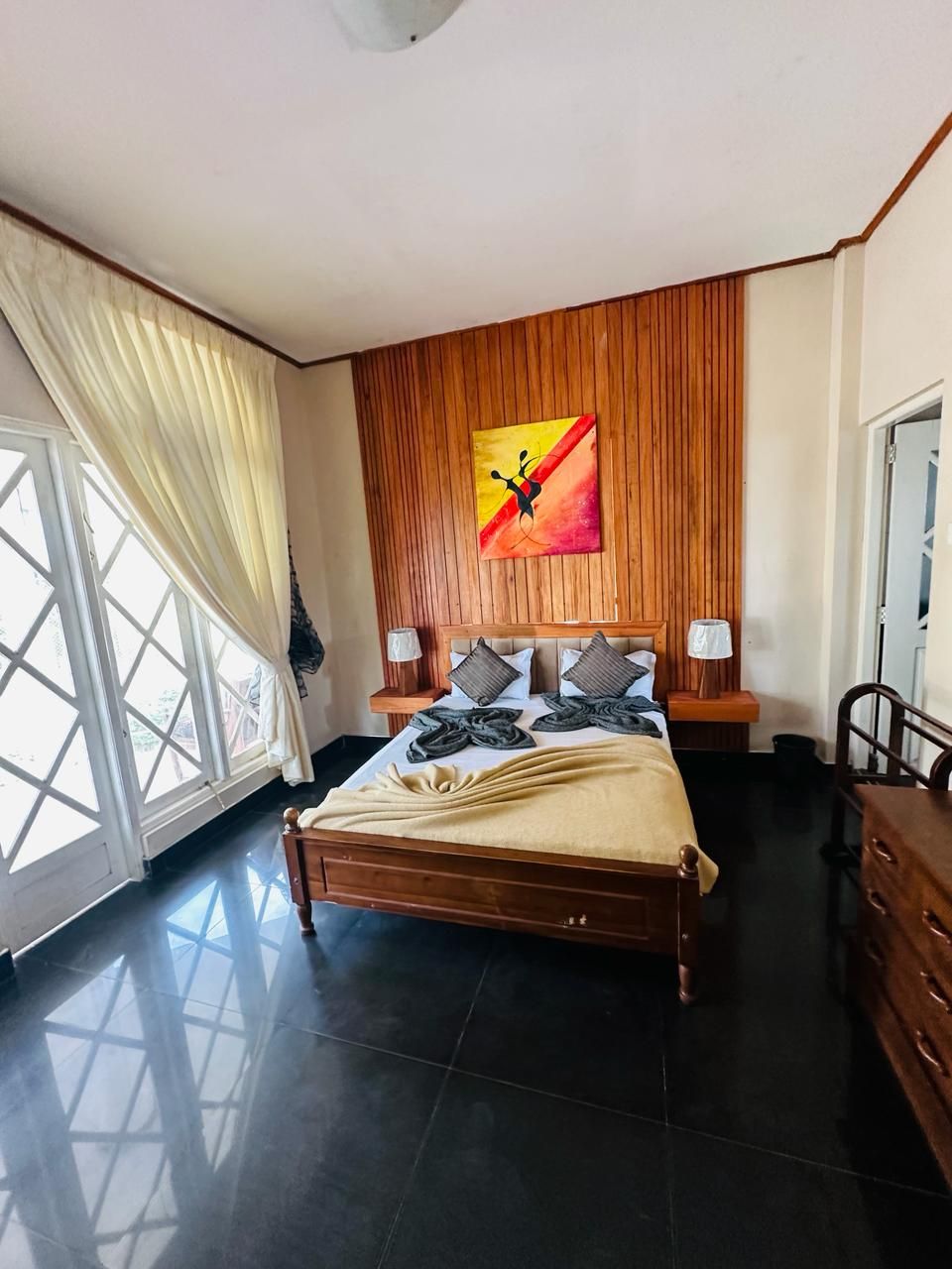 A bedroom with a wooden headboard, a bed with a beige blanket, two bedside lamps, and a large window with curtains.