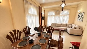 A dining area with a wooden table set for six, surrounded by chairs, a sofa, and a TV in the background. - MAPLES HOLIDAY RESORT (Nuwara Eliya)
