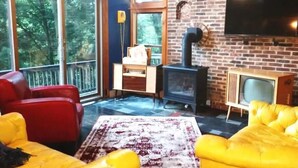 A living room with yellow sofas, a red chair, a fireplace, a TV, and large windows. - Red Bend cabin overlooking the river with hot tub. (Bellville)