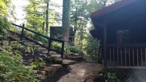 A wooden deck with a stone pathway leading to a covered area surrounded by trees. - Red Bend cabin overlooking the river with hot tub. (Bellville)