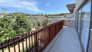 A balcony with a wooden railing, a glass door, and a view of a building and trees in the background. - Flat Ilê de Pipa Soé - By Almare (Tibau do Sul)