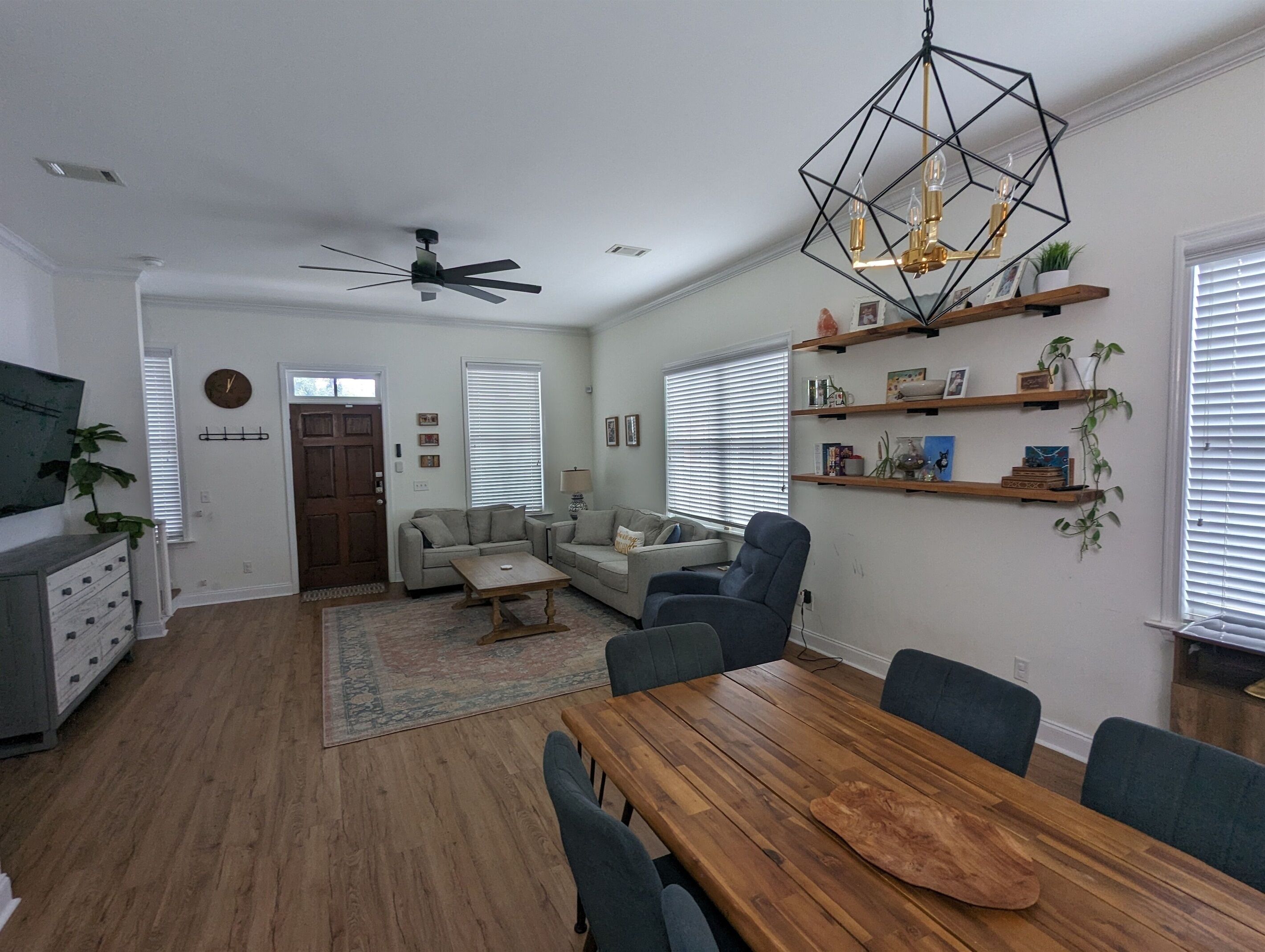 A modern living room with a wooden dining table, sofas, a ceiling fan, and a geometric pendant light.
