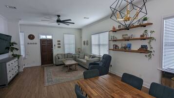 A modern living room with a wooden dining table, sofas, a ceiling fan, and a geometric pendant light.