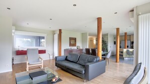 A modern living room with a grey sofa, a glass coffee table, and a wooden chair. There is a dining area with a table and chairs in the background.