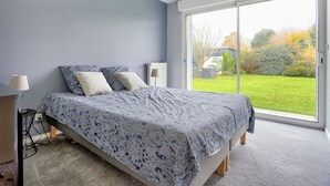 A bedroom with a bed, a nightstand, and a lamp. There is a large window with a view of a garden.