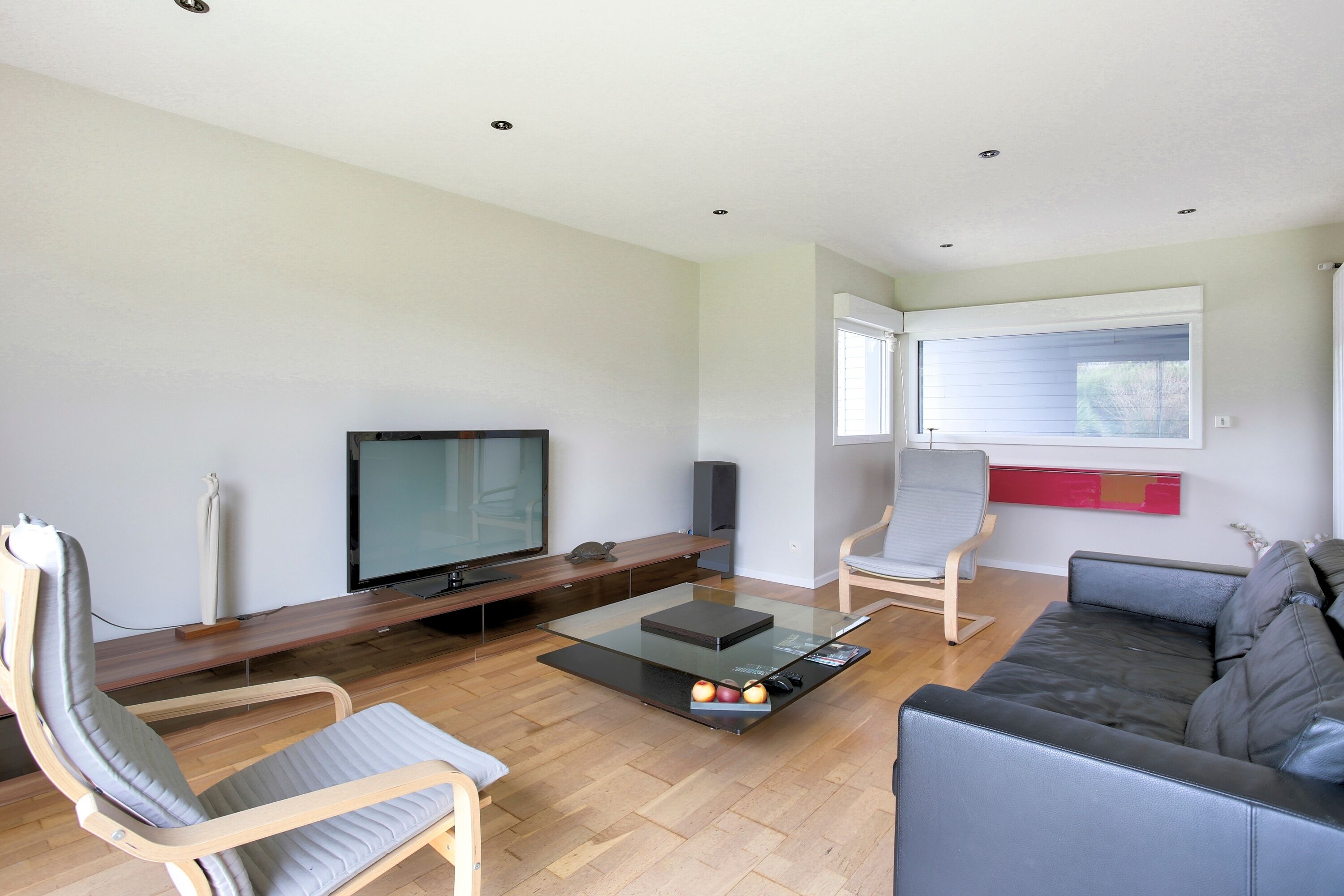A modern living room with a flat-screen TV, a glass coffee table with apples, a grey sofa, and a wooden bench.