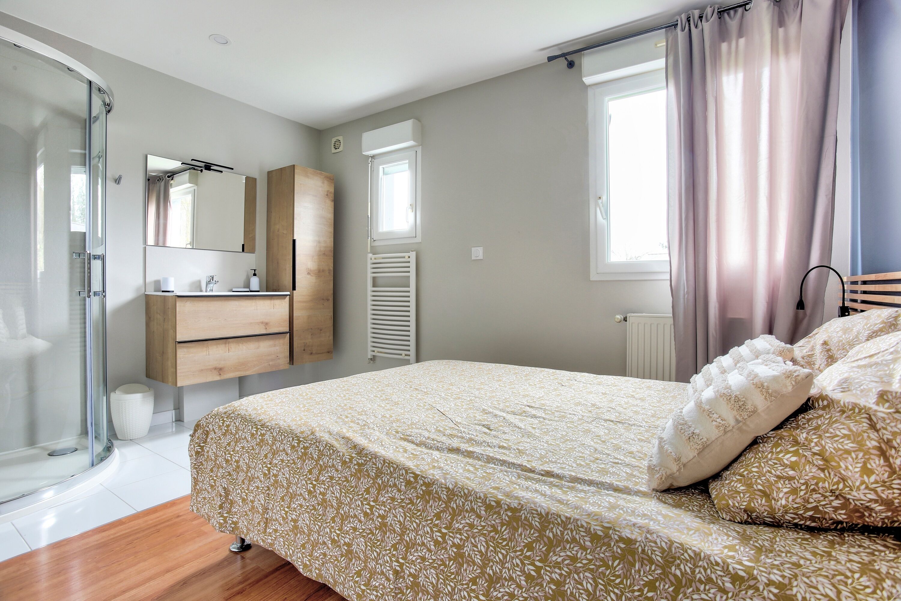 A bedroom with a bed, a shower, a vanity with a mirror, and a window with curtains.